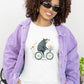 Woman wearing a white t-shirt with a cute bear riding a bicycle with a basket of flowers print